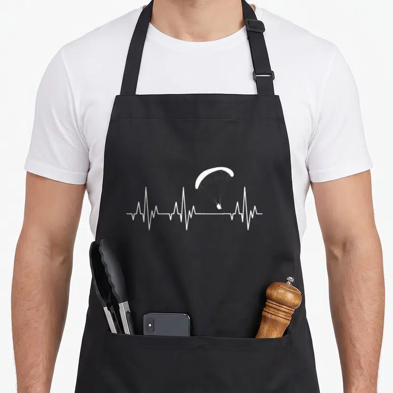Tablier de cuisine noir avec un battement de cœur blanc intégrant une silhouette de parapente en vol.