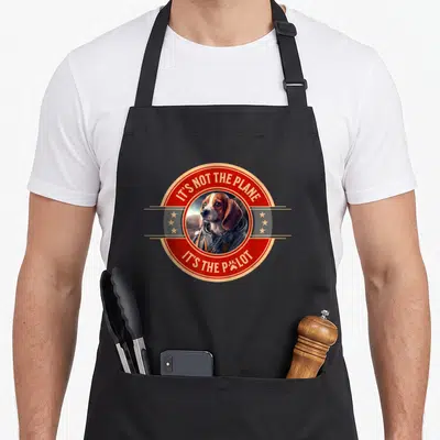 Tablier de cuisine noir avec un beagle en combinaison de pilote et texte It's not the plane it's the pilot dans un cercle rouge.