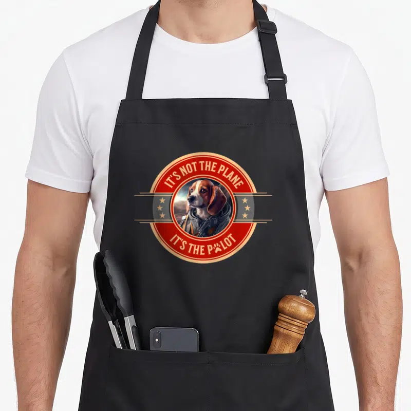 Tablier de cuisine noir avec un beagle en combinaison de pilote et texte It's not the plane it's the pilot dans un cercle rouge.