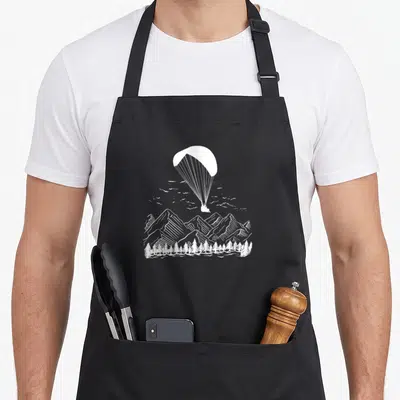 Tablier de cuisine noir avec illustration blanche d'un parapente au-dessus de sommets montagneux et d'une forêt de sapins.