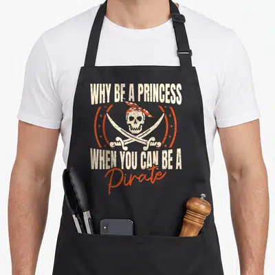 Tablier de cuisine noir avec crâne pirate, sabres croisés et texte Why Be A Princess When You Can Be A Pirate en orange et blanc.