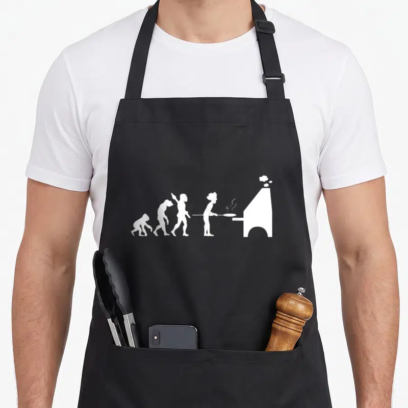 Tablier de cuisine noir avec une illustration blanche représentant l'évolution humaine se terminant par un pizzaiolo insérant une pizza dans un four à bois.