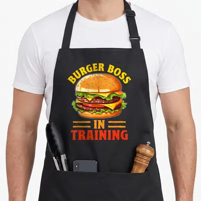 Tablier de cuisine noir avec illustration colorée d'un burger double et texte Burger Boss in Training en jaune et rouge.