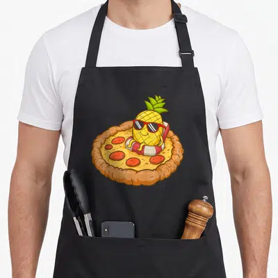 Tablier de cuisine noir avec illustration d'un ananas en lunettes de soleil dans une bouée sur une pizza hawaïenne.