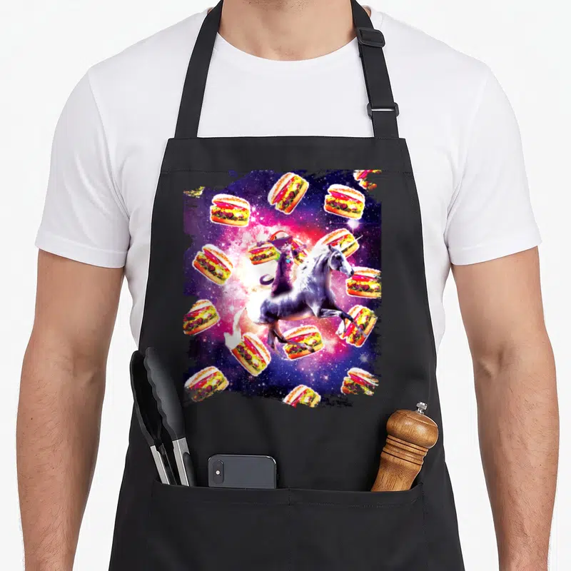 Tablier de cuisine noir avec un chat sur licorne volant parmi des burgers dans un univers spatial coloré.