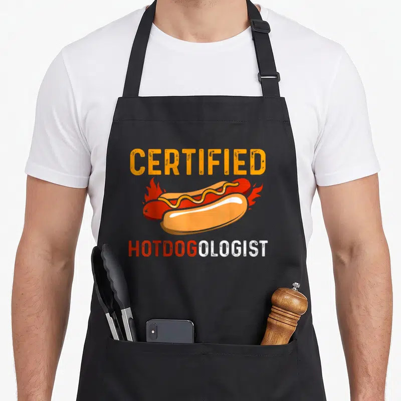Tablier de cuisine noir avec un hot dog enflammé et l'inscription Certified Hotdogologist en lettres orange et blanches.