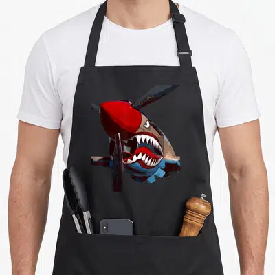 Tablier de cuisine noir avec illustration d'un avion de chasse P-40 Warhawk arbérant une tête de shark aux dents acérées sur le fuselage.