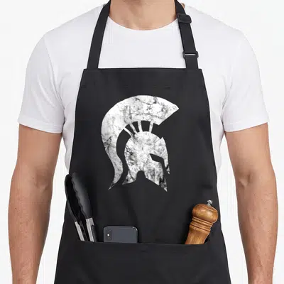 Tablier de cuisine noir avec illustration blanche texturée représentant un casque de gladiateur spartiate de style Leonida.