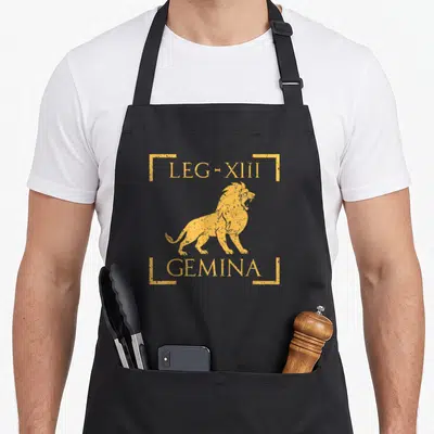 Tablier de cuisine noir avec un lion doré et les mots LEG-XIII GEMINA encadrés par des lignes jaunes.