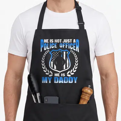 Tablier de cuisine noir avec inscription He is not just a police officer my daddy et silhouette d'agent sur insigne bleu.