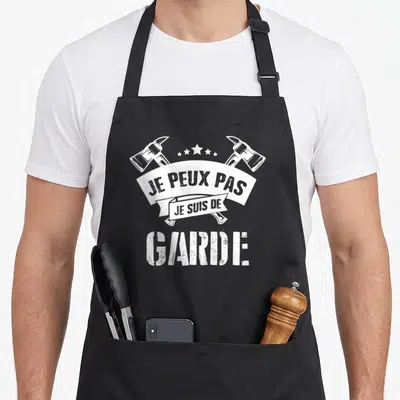 Tablier de cuisine noir avec inscription Je peux pas je suis de garde et deux haches croisées en blanc.