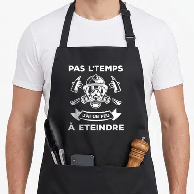 Tablier de cuisine noir avec casque de pompier, masque à gaz et haches croisées en blanc.