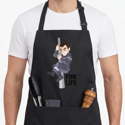 Tablier de cuisine noir avec illustration d'un sapeur pompier glissant sur un mât et le texte Fire Life en bas à droite.