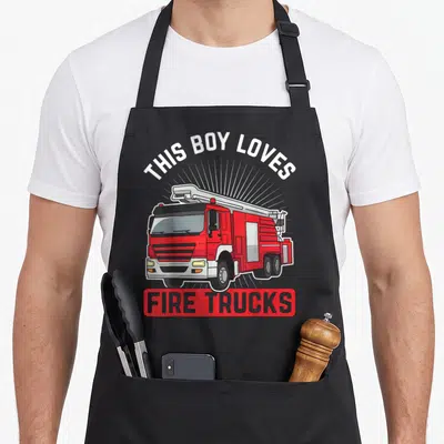 Tablier de cuisine noir avec illustration de camion de pompiers rouge et texte This Boy Loves Fire Trucks en anglais.