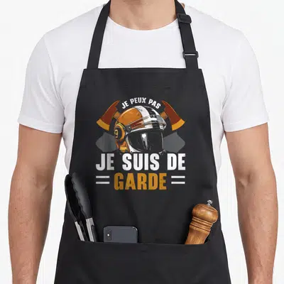 Tablier de cuisine noir avec casque de pompier orange et inscription Je peux pas je suis de garde en blanc et jaune.