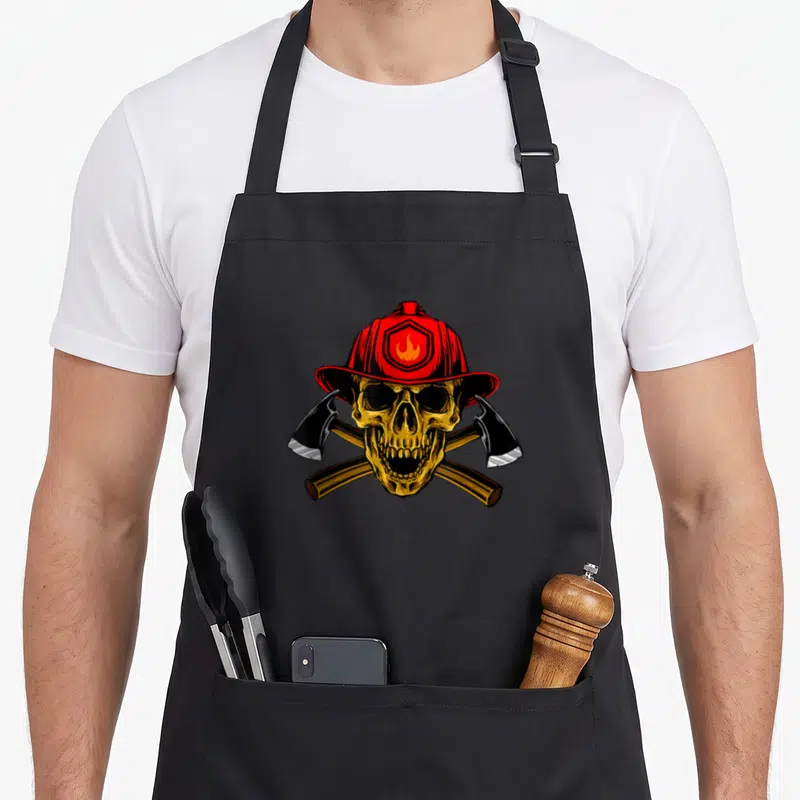 Tablier de cuisine noir avec un crâne doré portant un casque de pompier rouge et deux haches croisées en arrière-plan.