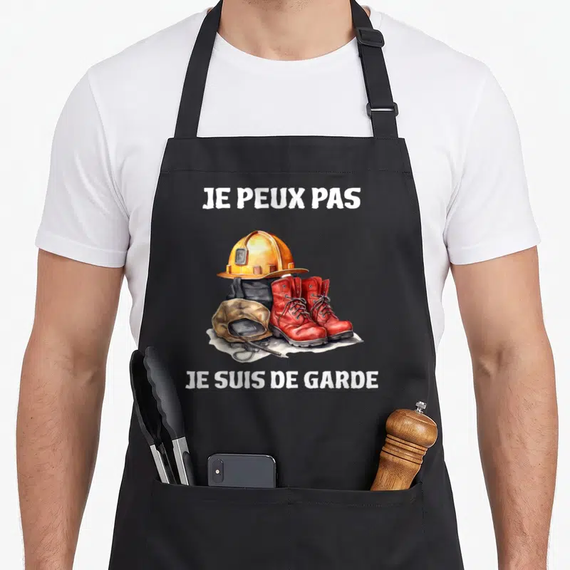 Tablier de cuisine noir avec casque orange, bottes rouges et texte Je peux pas je suis de garde imprimé en blanc.