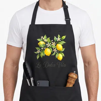 Tablier de cuisine noir avec branchage de citrons jaunes, feuilles vertes, fleurs blanches et texte La Dolce Vita en bas.