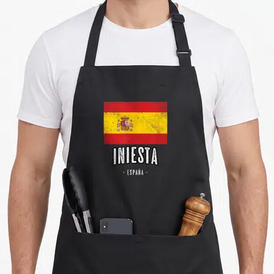 Tablier de cuisine noir avec le drapeau espagnol vintage et le texte Iniesta España imprimé en blanc au centre.
