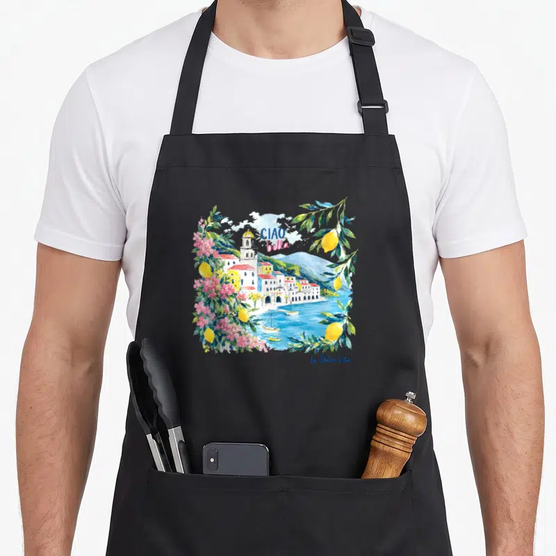 Tablier de cuisine noir avec une illustration colorée représentant un village côtier italien encadré de citrons et de fleurs roses.