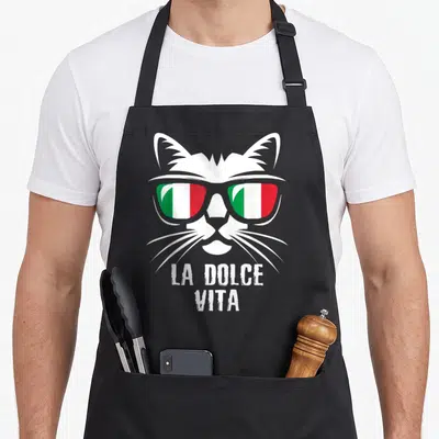 Tablier de cuisine noir avec un chat blanc stylisé portant des lunettes aux couleurs du drapeau italien et le texte La Dolce Vita en dessous.