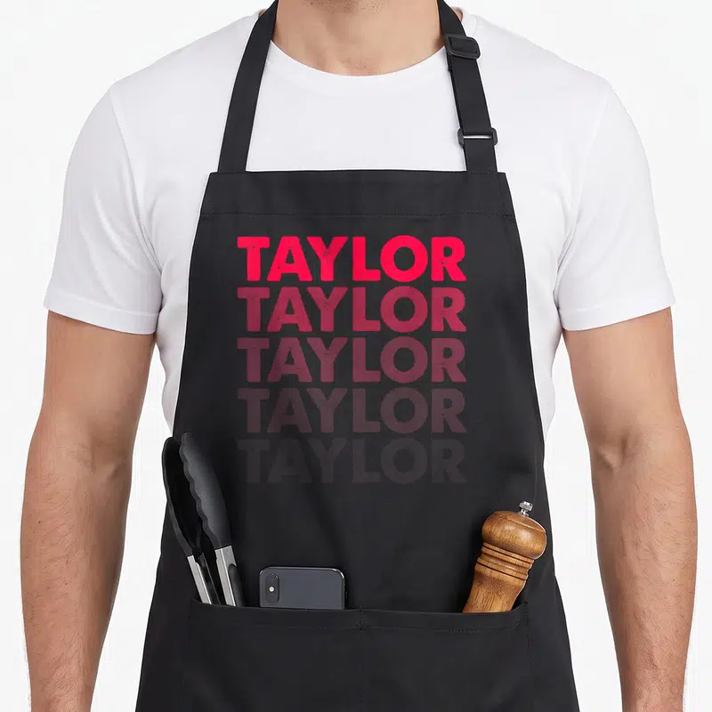 Tablier de cuisine noir avec le prénom TAYLOR imprimé en rose vif et dégradé sur cinq lignes verticales.