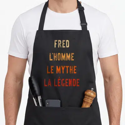 Tablier de cuisine noir avec inscription Fred lhomme le mythe la légende en lettres orange et rouge dégradé.