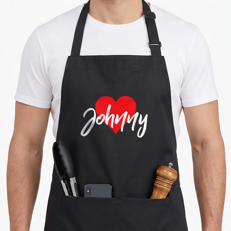 Tablier de cuisine noir avec un cœur rouge et le prénom Johnny écrit en blanc sur la poitrine.