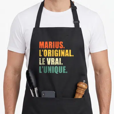Tablier de cuisine noir avec les mots Marius loriginal le vrai et lunique imprimés en couleurs vives sur le devant.