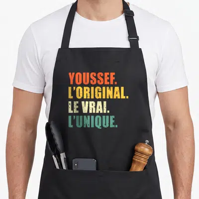 Tablier de cuisine noir avec les mots Youssef L'original Le Vrai L'unique imprimés en lettres colorées.