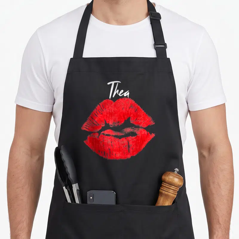 Tablier de cuisine noir avec le prénom Thea en blanc au-dessus d'une trace de rouge à lèvres rouge vif.