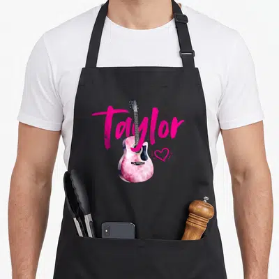 Tablier de cuisine noir avec une guitare rose tie-dye, le nom Taylor en rose et un petit cœur stylisé.