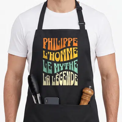 Tablier de cuisine noir avec inscription l'homme le mythe la légende en lettres ondulées orange jaune vert et crème.