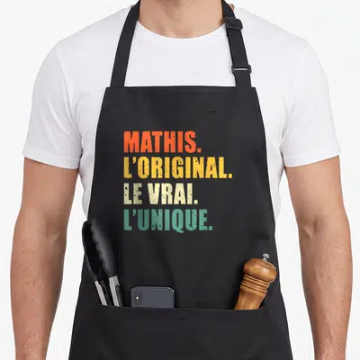 Tablier de cuisine noir avec le texte coloré Mathis l'original le vrai l'unique imprimé en grosses lettres capitales au centre du vêtement.
