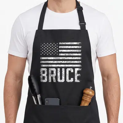 Tablier de cuisine noir avec l'inscription BRUCE en blanc et un drapeau américain texturé au-dessus.