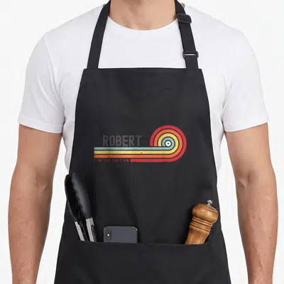 Tablier de cuisine noir avec le prénom Robert et un motif circulaire multicolore style rétro sur fond uni.