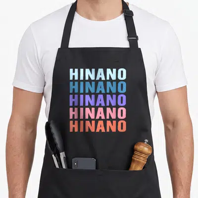 Tablier de cuisine noir avec le prénom HINANO imprimé en lettres colorées superposées verticalement.