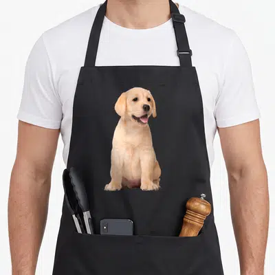 Tablier de cuisine noir avec un chiot Golden Retriever assis, pattes avant visibles et langue rose sortant joyeusement.