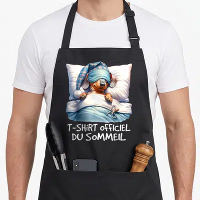 Tablier de cuisine noir avec un teckel dormant sous une couverte bleue, portant un masque et une bonnette à carreaux.