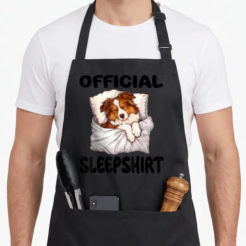 Tablier de cuisine noir avec illustration d'un Berger australien dormant sur une oreiller et le texte Official Sleepshirt imprimé en noir.