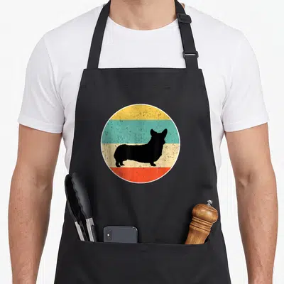 Tablier de cuisine noir avec silhouette de chien corgi sur fond circulaire multicolore vintage.