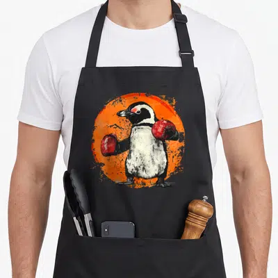 Tablier de cuisine noir avec illustration d'un pingouin portant des gants de boxe rouges sur fond orange texturé.