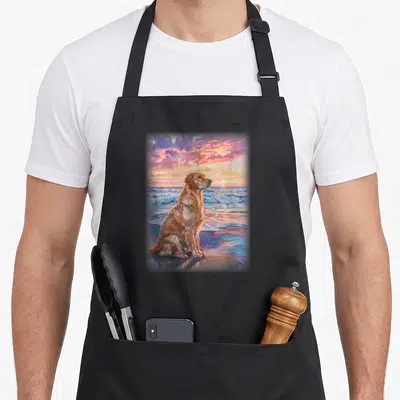 Tablier de cuisine noir avec illustration d'un Golden Labrador assis sur le sable face à l'océan sous un ciel rose et orange.