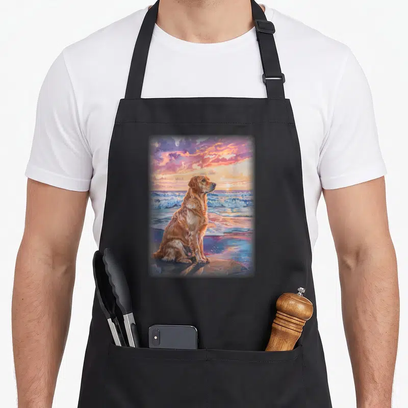 Tablier de cuisine noir avec illustration d'un Golden Labrador assis sur le sable face à l'océan sous un ciel rose et orange.