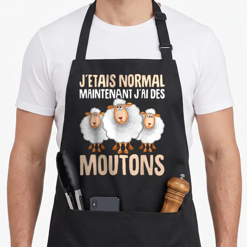 Tablier de cuisine noir avec trois moutons blancs et le texte J'étais normal maintenant j'ai des moutons imprimé en lettres capitales.
