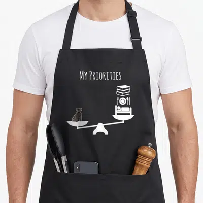 Tablier de cuisine noir avec une balance montrant un chien plus lourd que des livres et de la nourriture.
