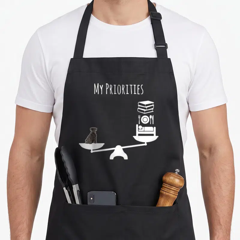 Tablier de cuisine noir avec une balance montrant un chien plus lourd que des livres et de la nourriture.