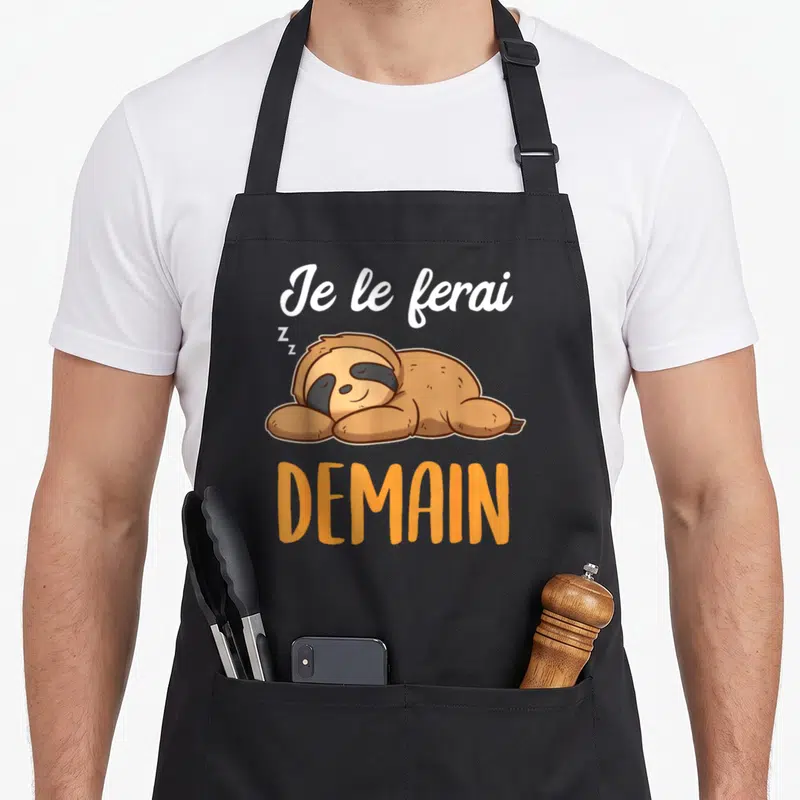 Tablier de cuisine noir avec un sloth marron dormant et le texte Je le ferai demain en orange et blanc.