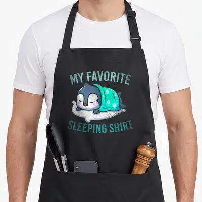 Tablier de cuisine noir avec illustration d'un pingouin dormant sous une couverture turquoise et le texte My Favorite Sleeping Shirt.
