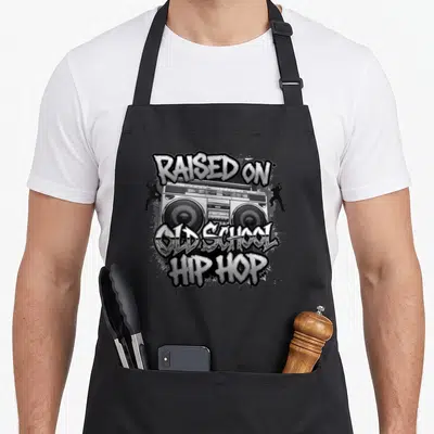 Tablier de cuisine noir avec boombox, silhouettes de danseurs et texte Raised on Old School Hip Hop en style graffiti.